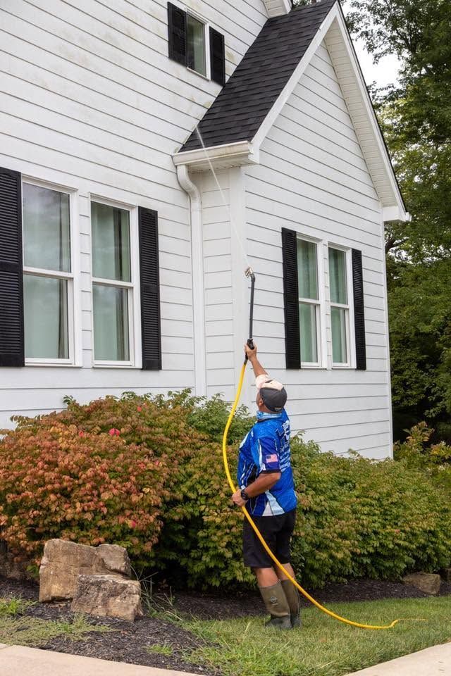 Person using a hose to spray water against a white house exterior and bushes.