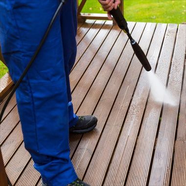 Person pressure washing a wooden deck with a black hose and white spray.