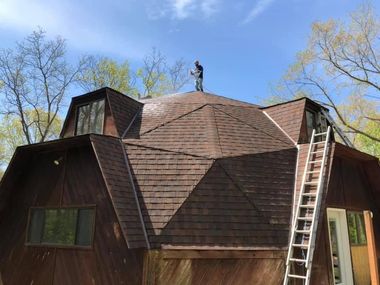 A wooden dome house with a ladder leaning against it and a person on the roof, under a blue sky.