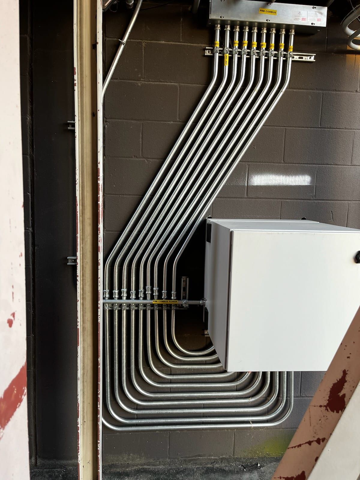 Electrical conduit pipes on a brick wall connect to a white box and top panel, with a vertical support on the left.