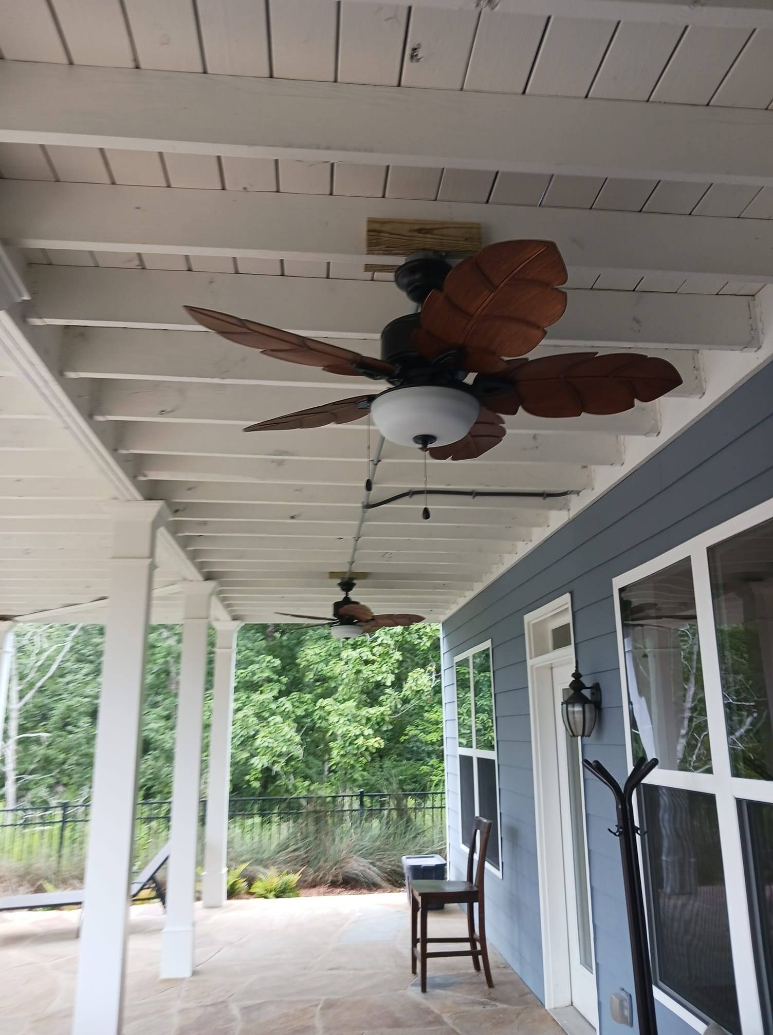 Two ceiling fans under a covered porch with white beams, a blue wall, and a stone floor.