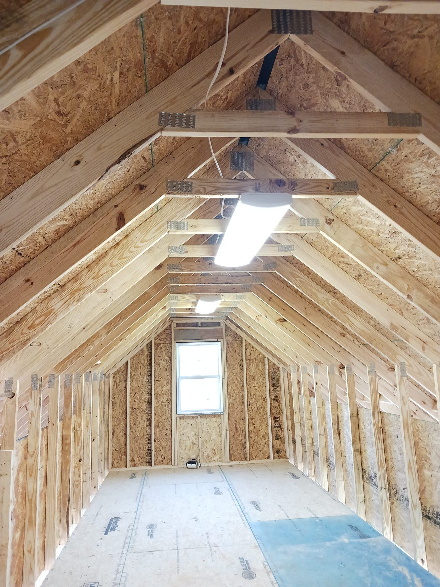 Interior view of an unfinished attic with exposed wooden beams and rafters, a window, and a fluorescent light fixture.