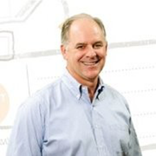Man with light skin smiles, wearing a light blue button-down shirt. Background features a map or diagram.