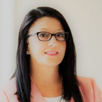 Woman with dark hair and glasses smiles, wearing a pink blazer.