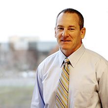 Man in light blue shirt and striped tie, smiling indoors, blurred background.