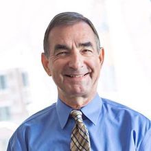 Man in blue shirt and tie smiling, inside with blurred background.