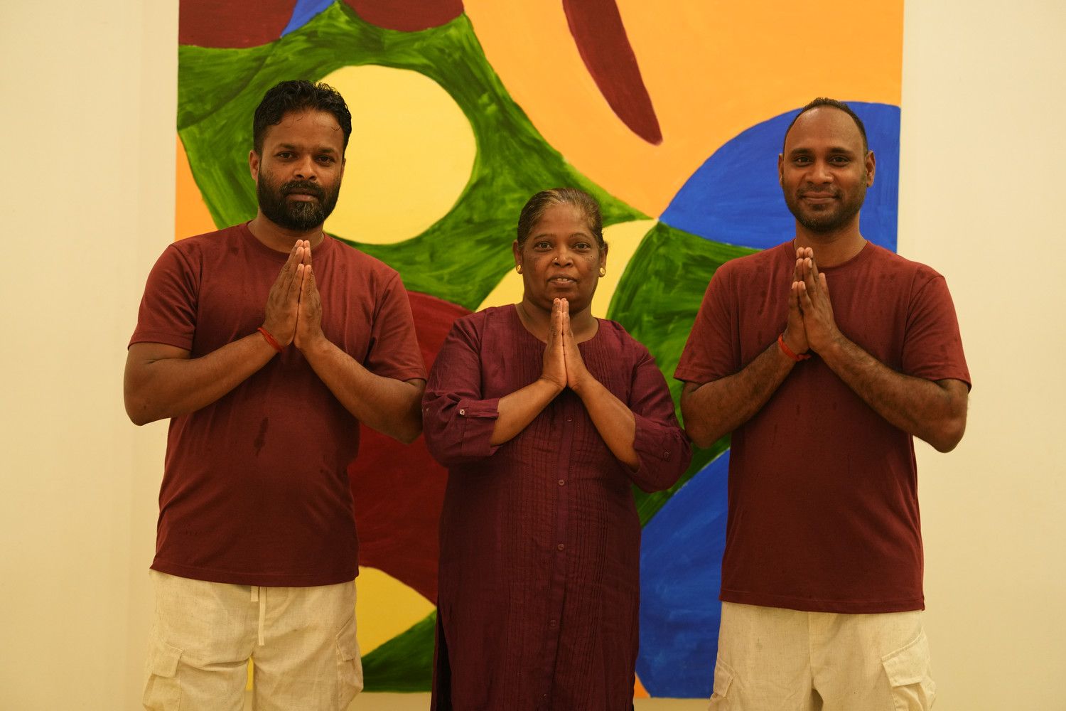 Three people with hands together in prayer pose, in front of a colorful abstract painting.
