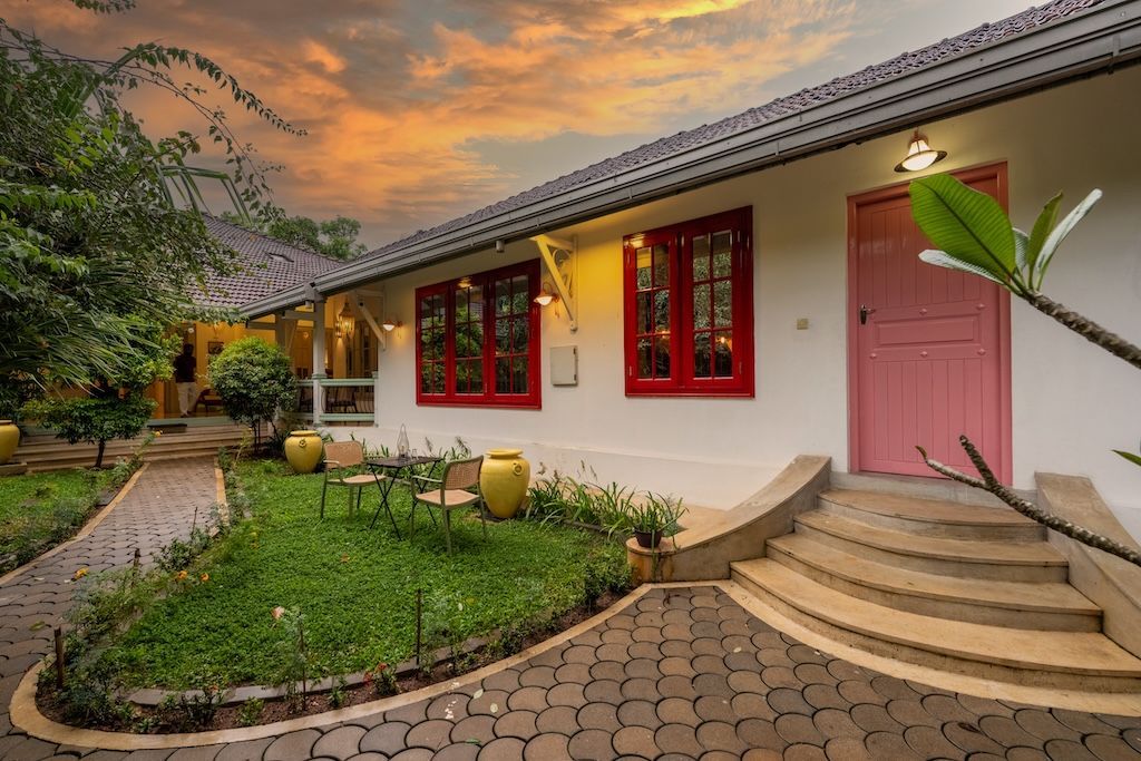 White building with red-framed windows and a pink door. Cobblestone path, green lawn, sunset.