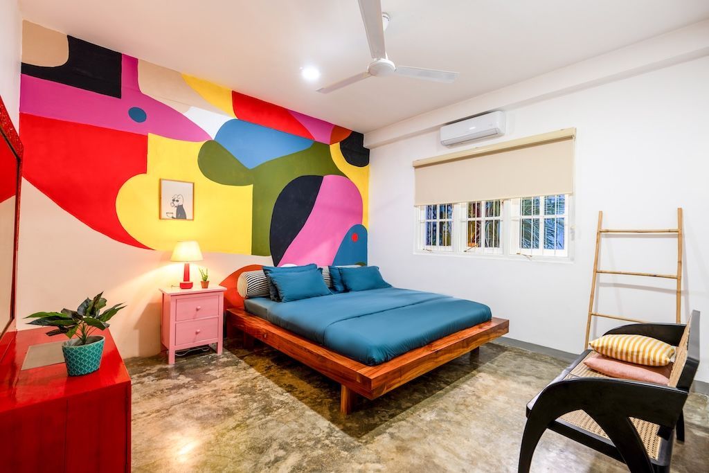 Bedroom with colorful mural wall, wood bed, pink nightstand, red dresser, and black chair.