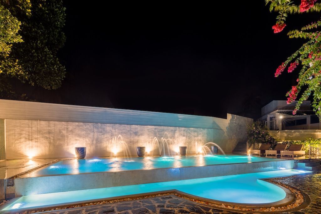 Night view of a swimming pool with water fountains illuminated. Bougainvillea and building in the background.