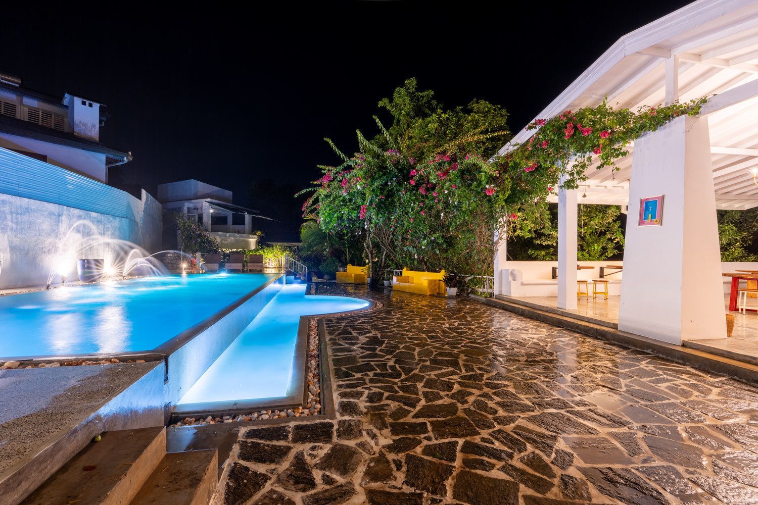 Nighttime view of a lit pool with a waterfall, stone patio, and covered patio with seating.