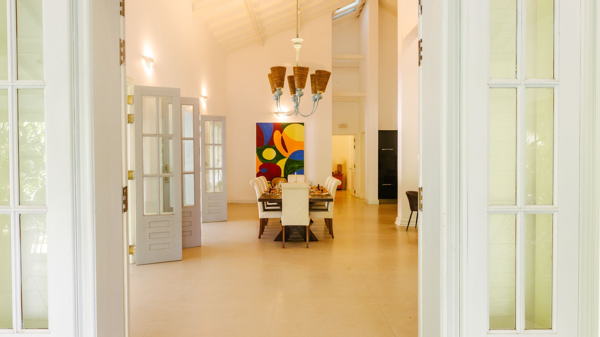 Open white doors frame a bright room with a dining table, artwork, and a chandelier.