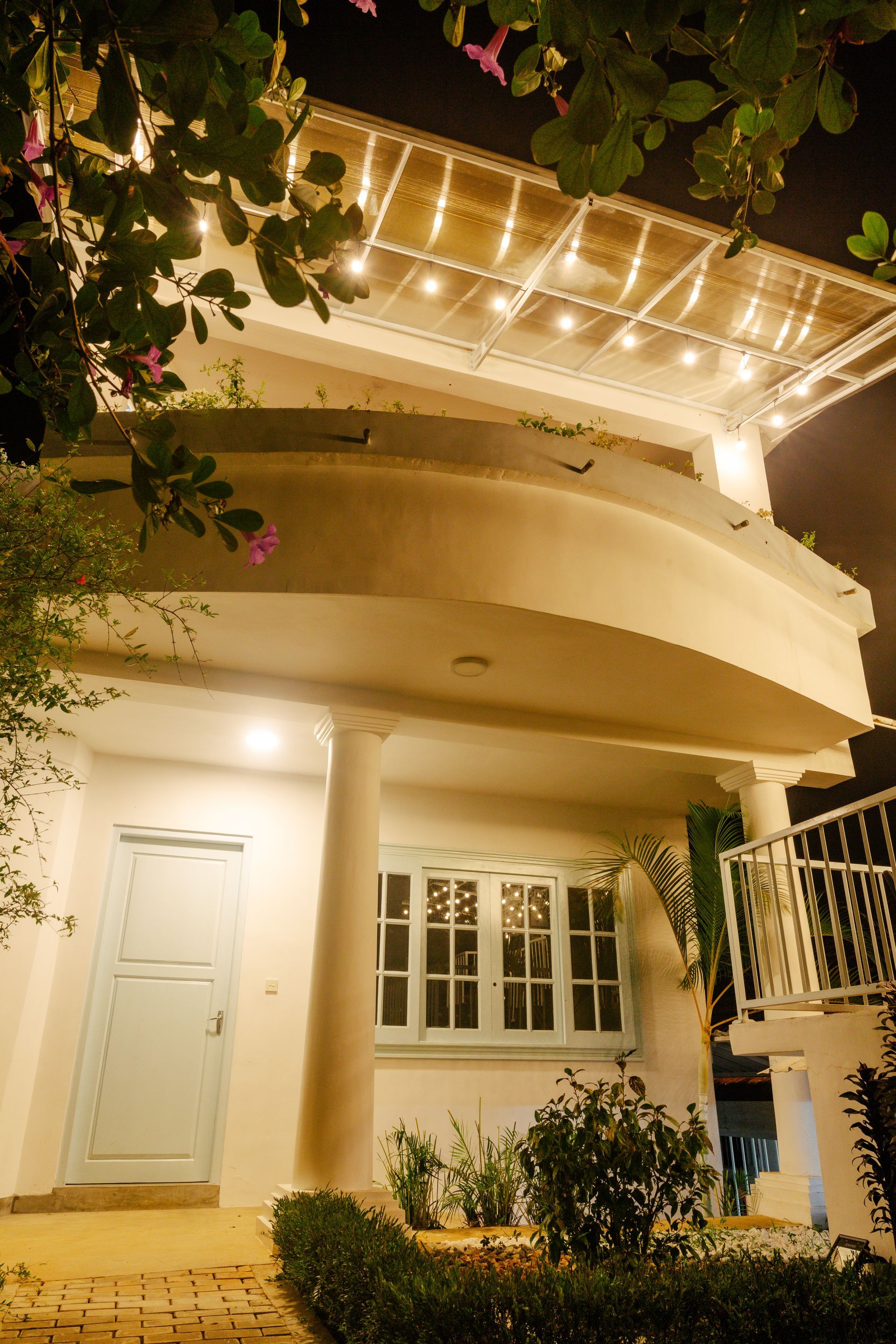 White house exterior with illuminated facade at night; curved balcony, blue door, and cascading vines.