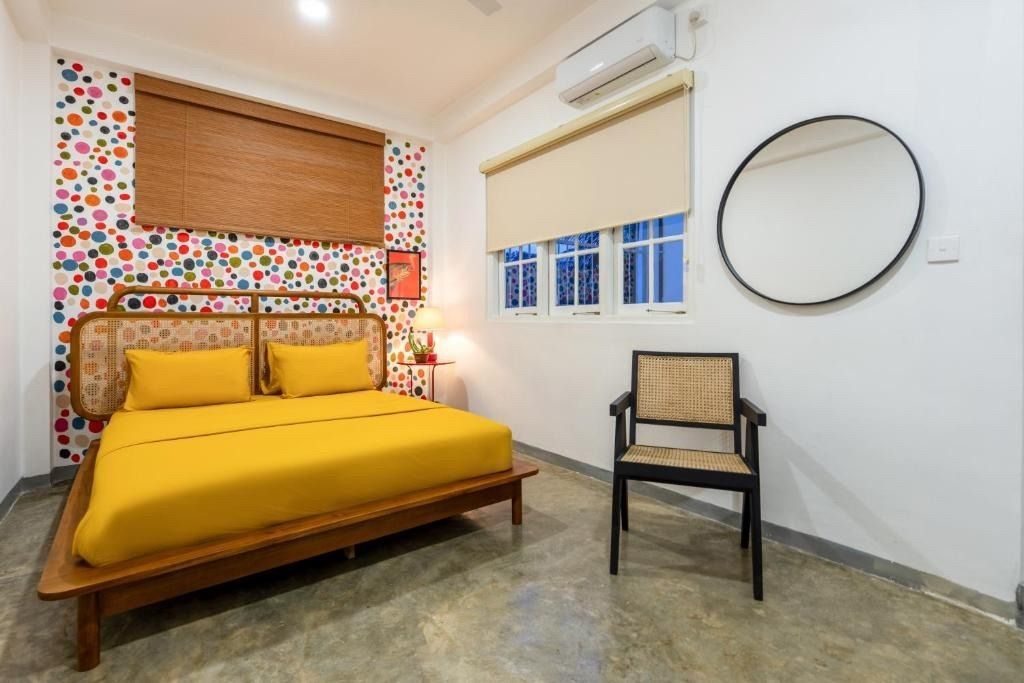 Bedroom with a yellow bed, patterned wall, chair, mirror, and a window with a beige roller blind.