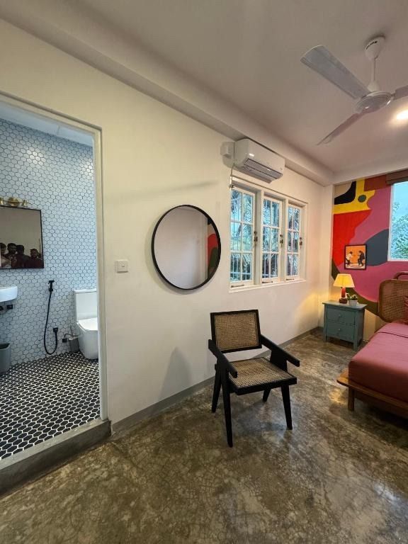 Bedroom with bathroom entrance, round mirror, chair, and bed; colorful artwork and window.