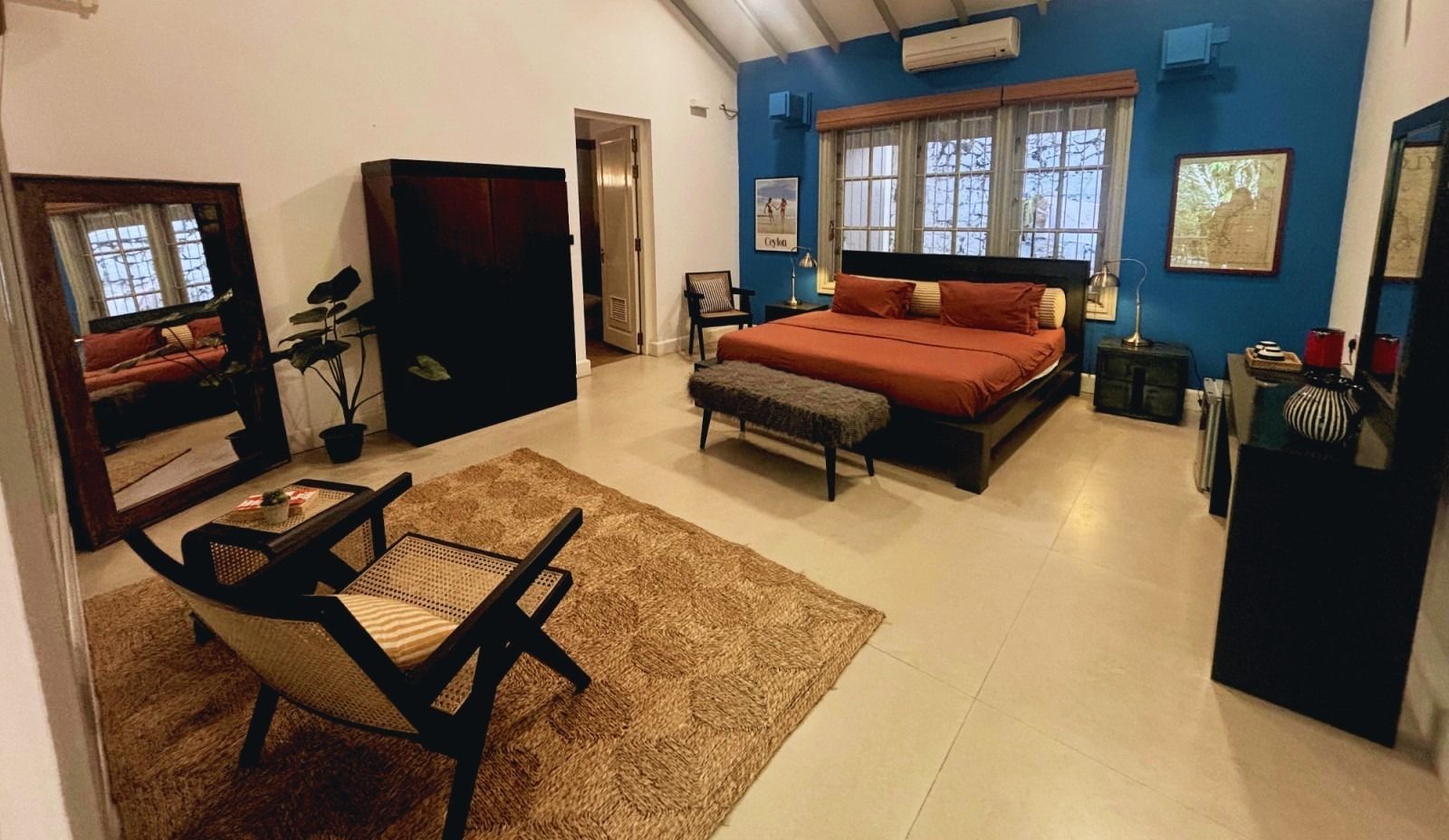 Spacious bedroom with a king-sized bed, large mirror, and seating area on a plush rug. Blue walls and natural light.