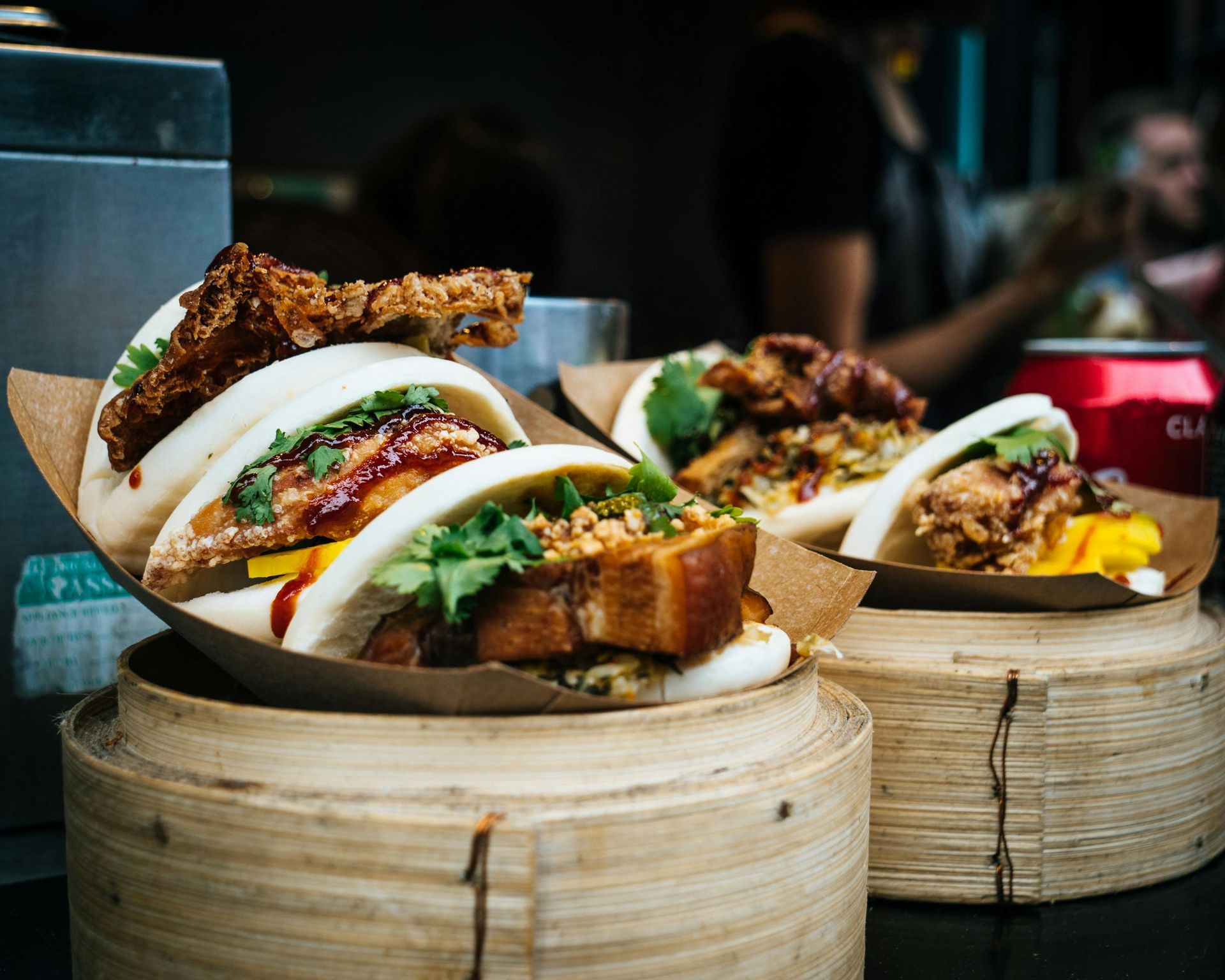 Three bao buns topped with meat, cilantro, and garnish, presented in wooden bamboo steamers at a food stall.