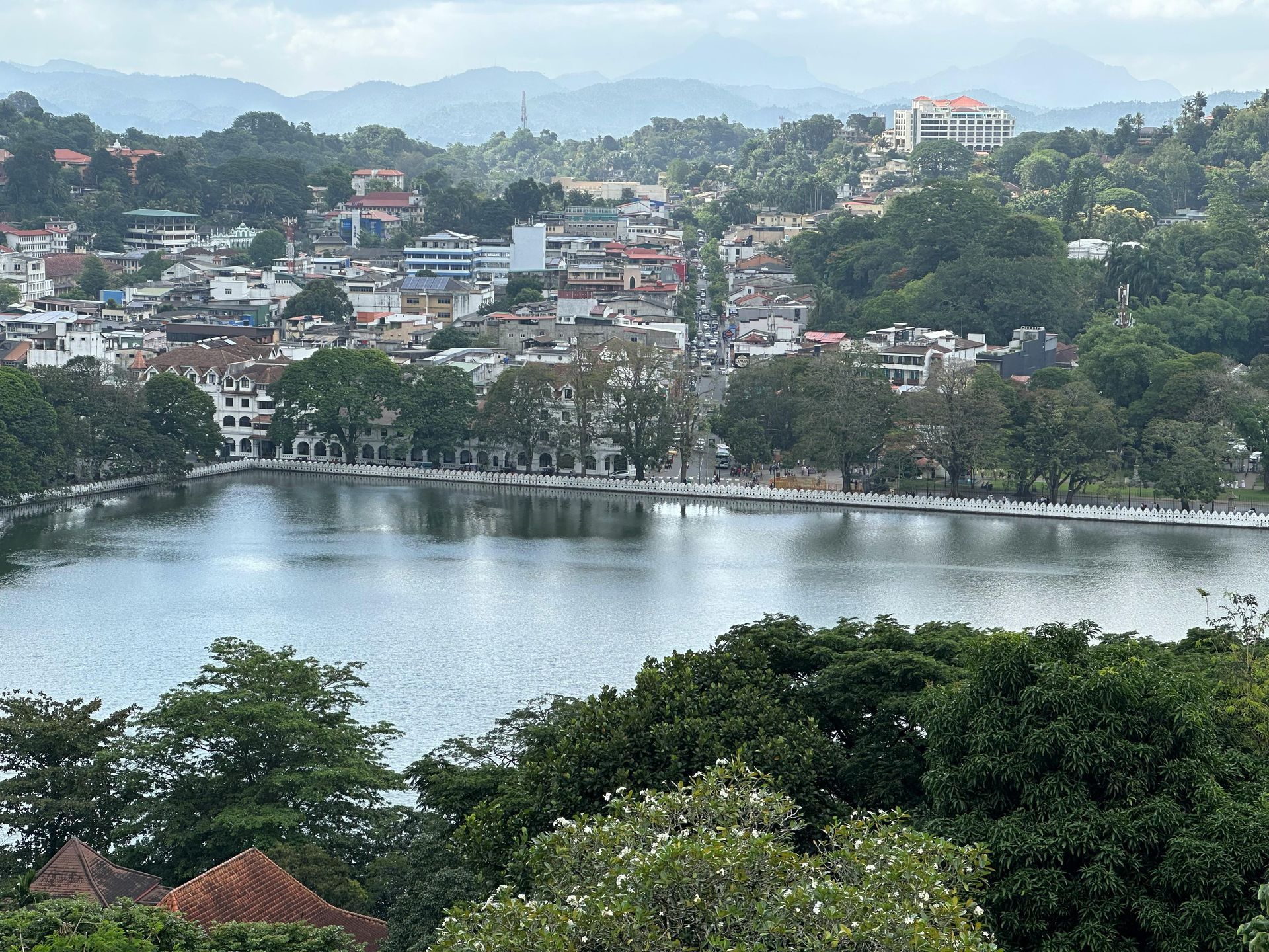 Cityscape with a lake in the foreground, buildings and greenery spread across the scene, and mountains in the distance.