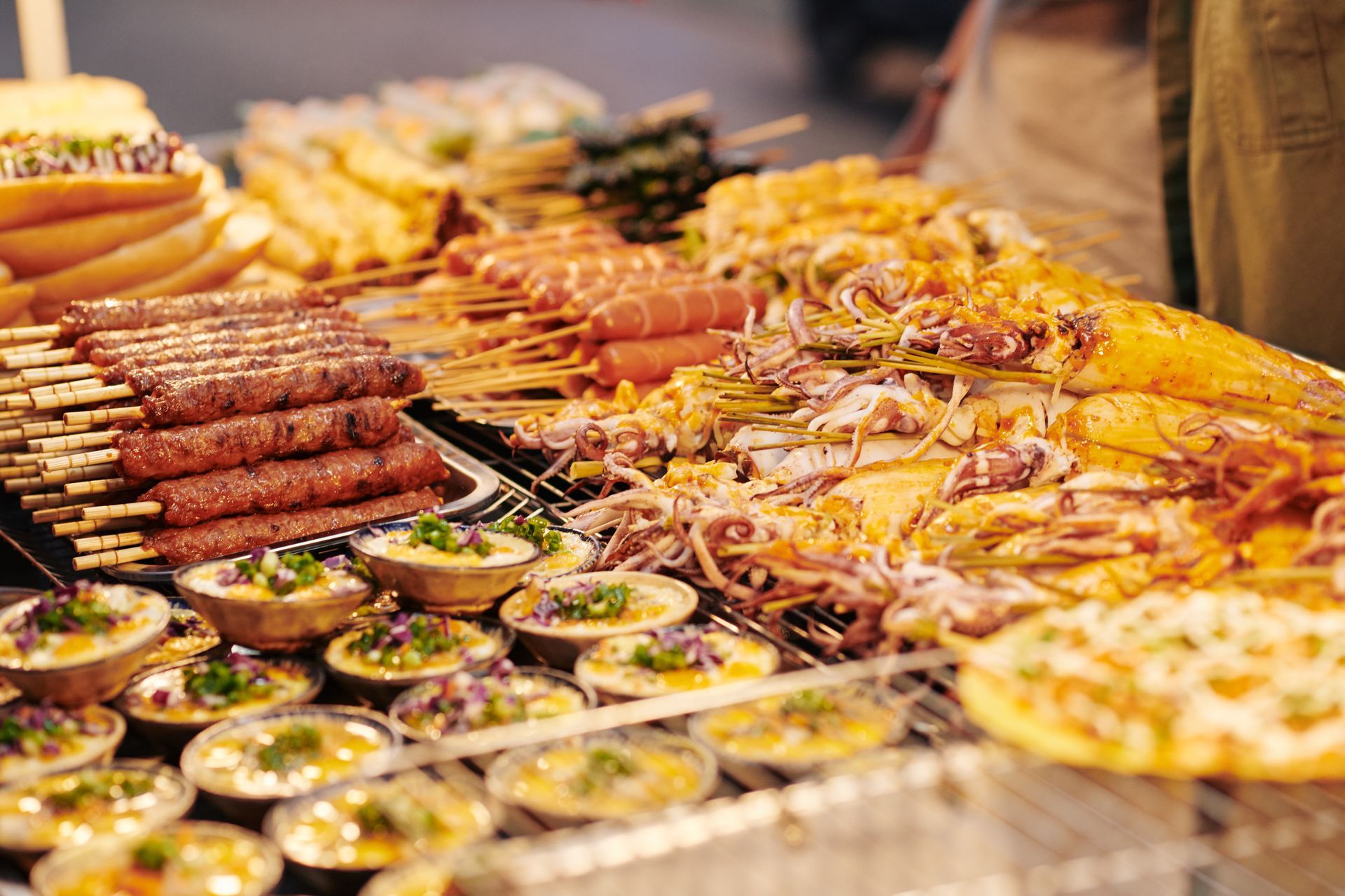 A vibrant street food stall featuring skewers, grilled meats, seafood, and small bowls of cooked appetizers.