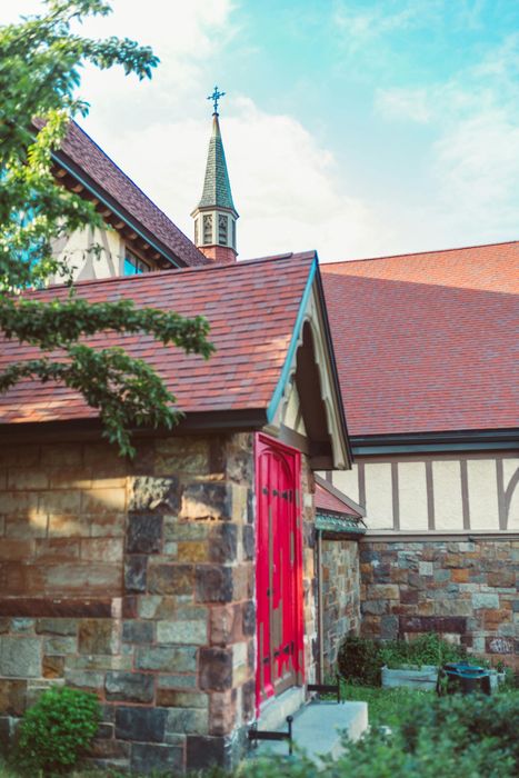 Red door of stone church with steeple, red tile roof, and green shrubbery.