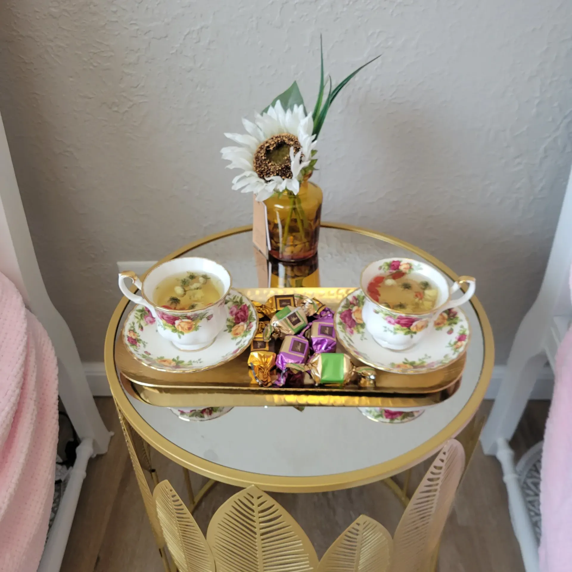 Two teacups, candies, and a flower vase on a gold tray atop a small mirrored table.