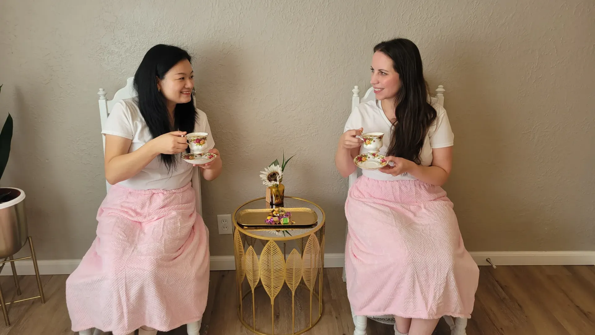 Two women in matching pink skirts and white tops, seated, talking, holding mugs.