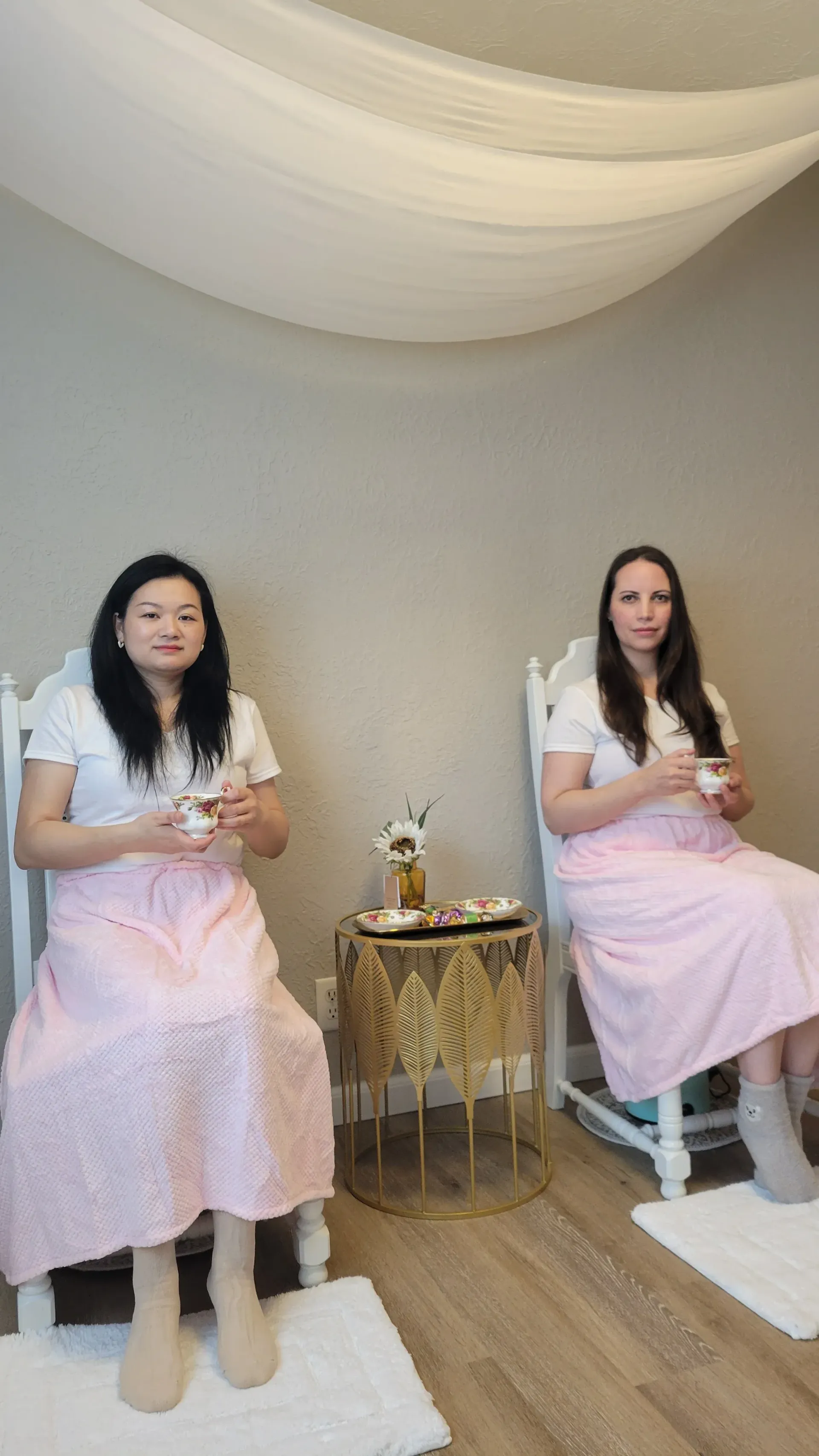 Two women in matching pink skirts and white tops sit on white chairs, holding cups. Neutral setting.