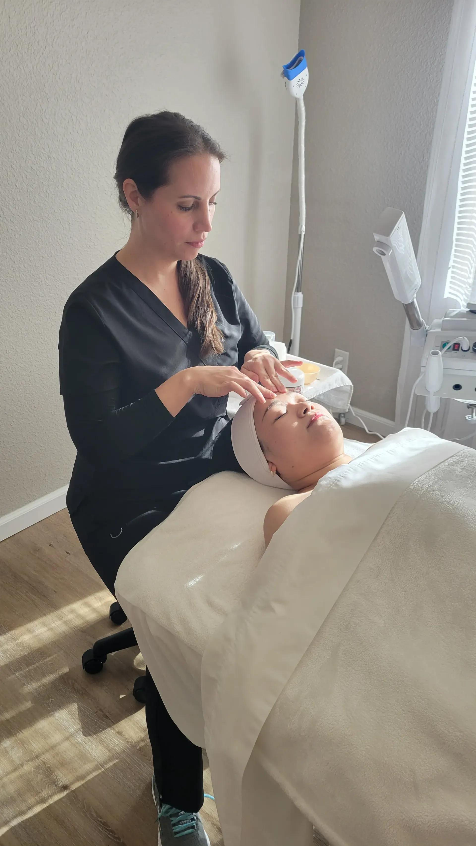 Esthetician giving a facial to a client lying on a spa bed in a treatment room.