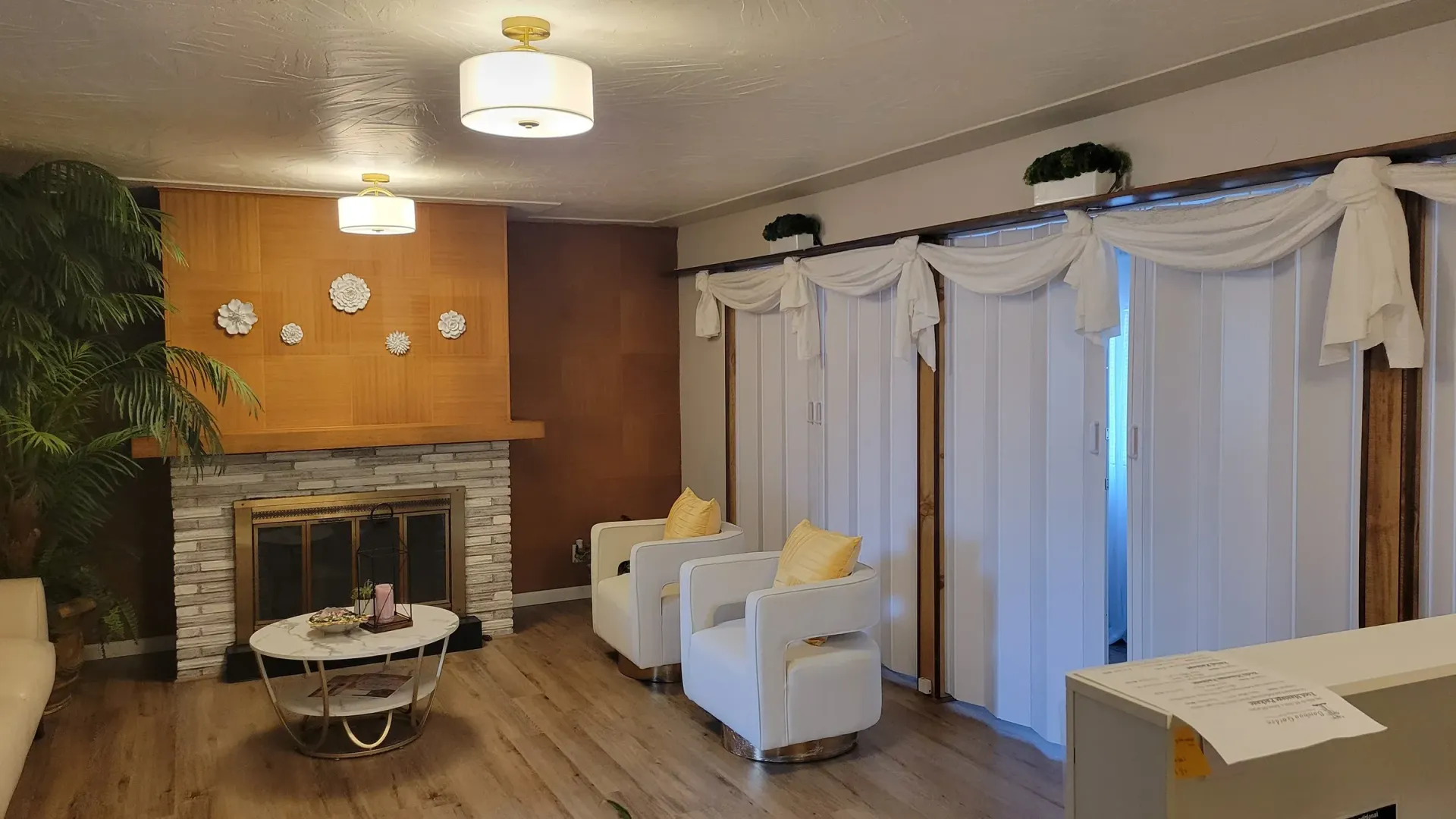Living room with fireplace, white curtains, chairs, and coffee table. Wooden paneling and hardwood floors.