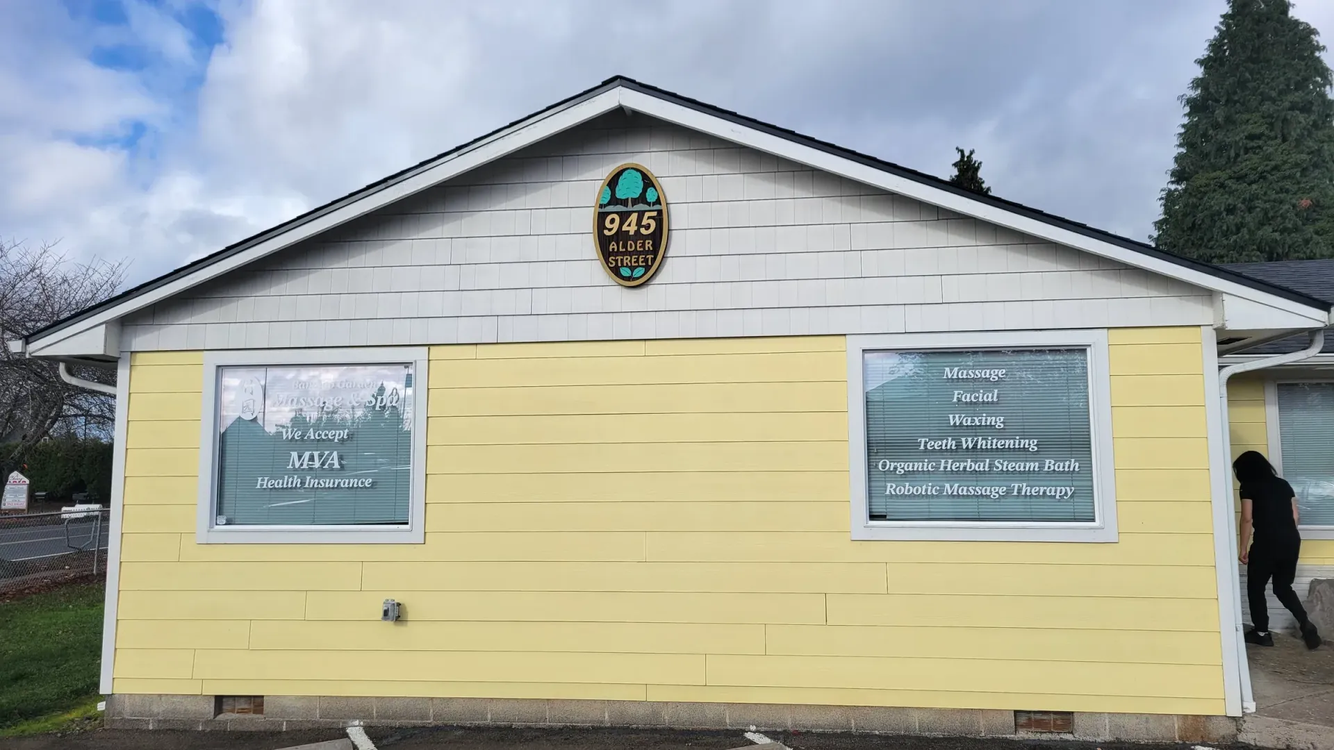 Yellow house with two windows, a sign, and a person walking on the right. Cloudy sky.