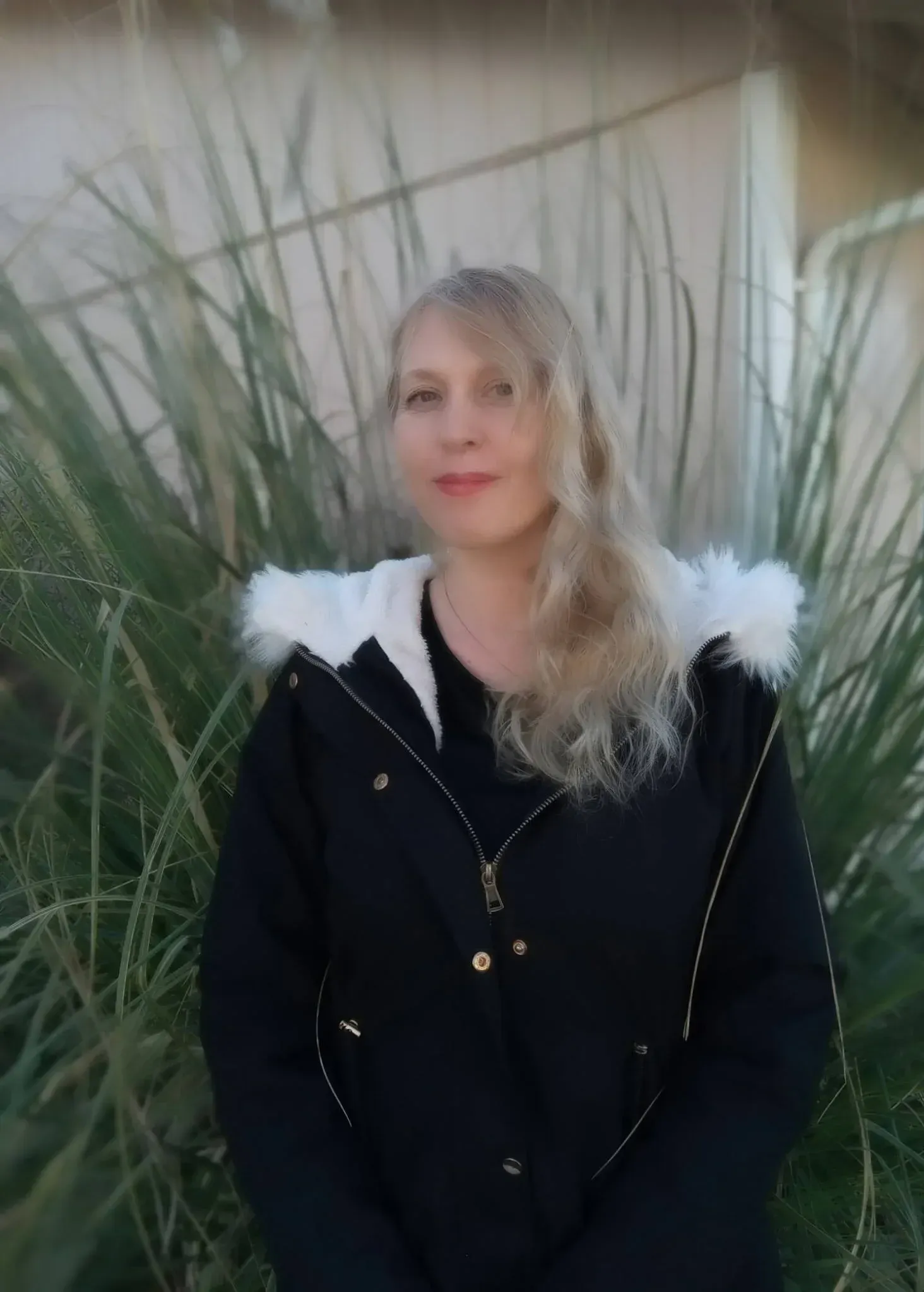Woman with long blonde hair wearing a black jacket with a white fur-trimmed hood, standing outdoors near green plants.