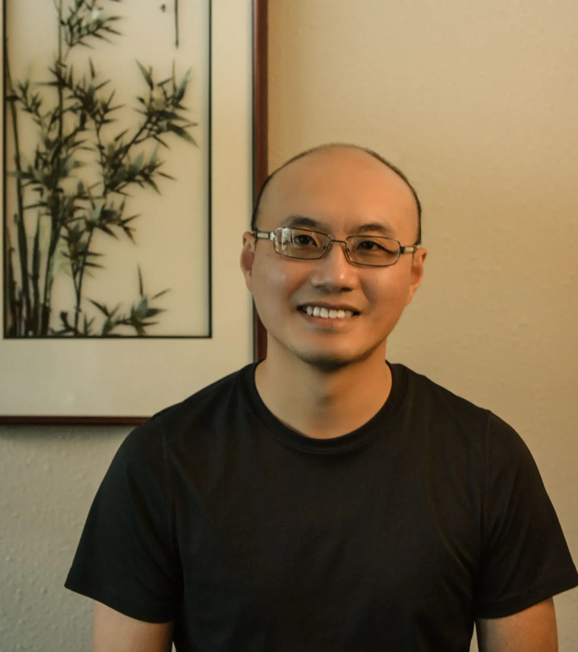 Man with glasses smiling in front of a framed bamboo art piece. Wearing a black shirt.