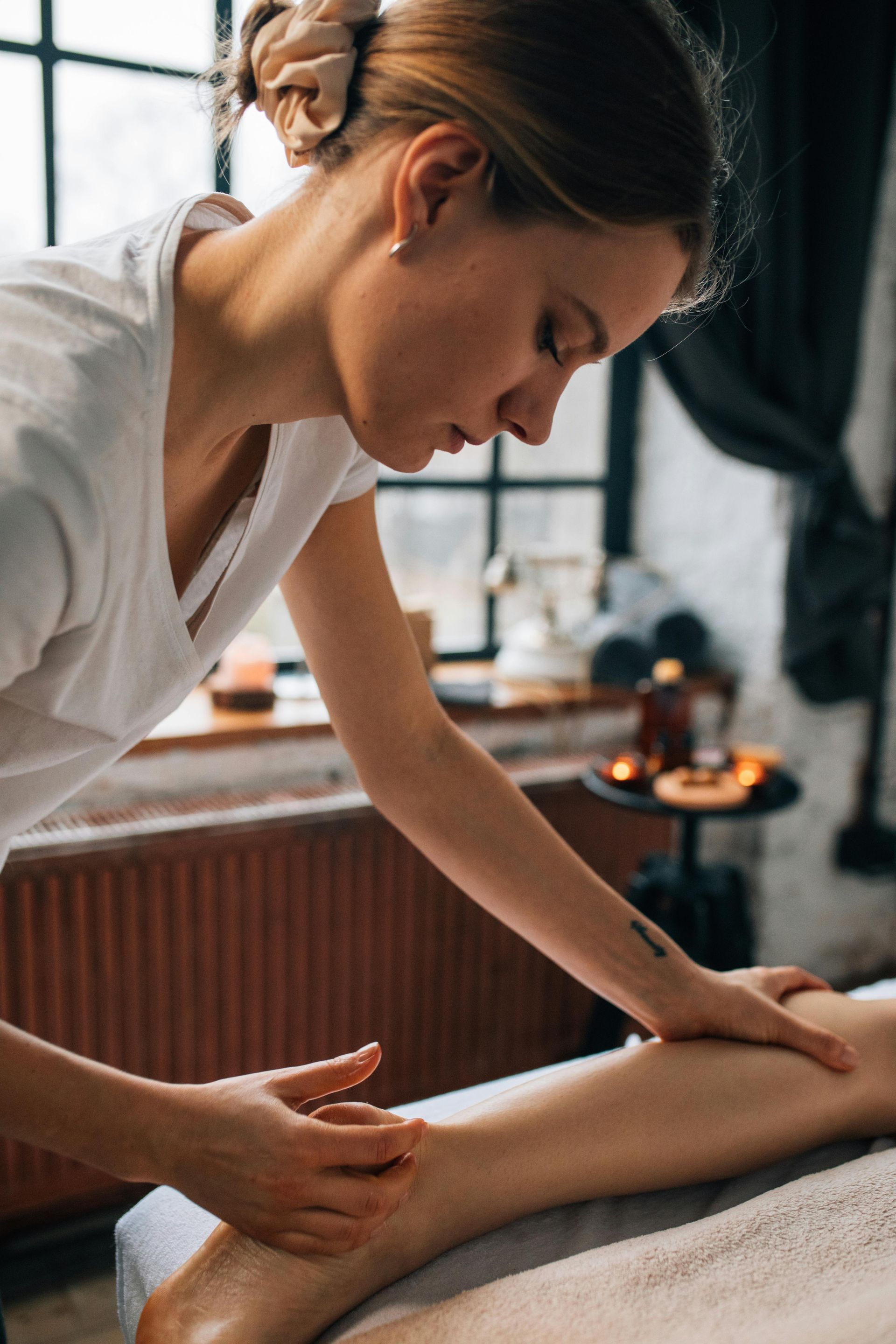 Woman massaging a leg in a spa setting; window in the background, warm tones.