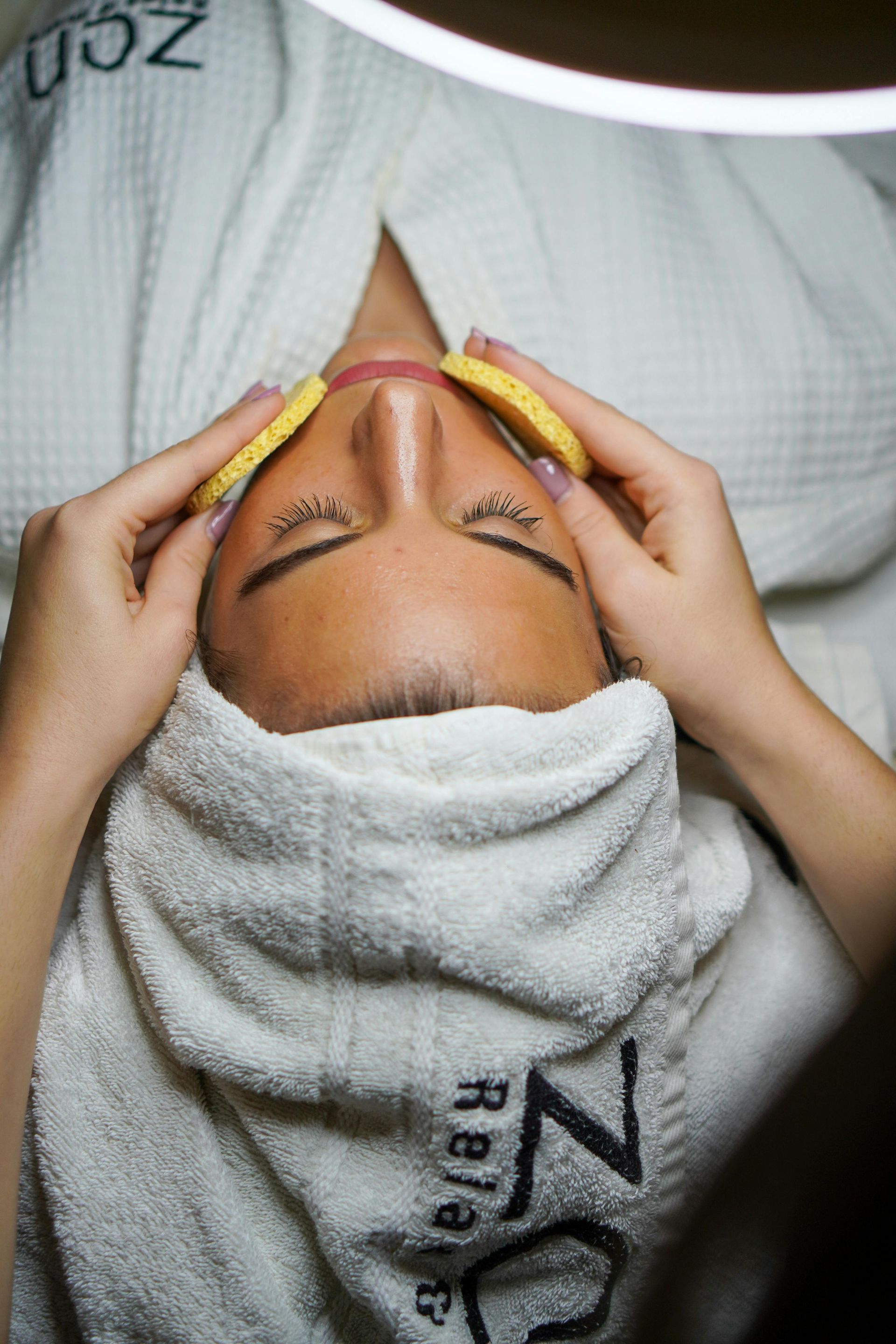 Woman receiving facial treatment, covered with a towel and robe, eyes closed.