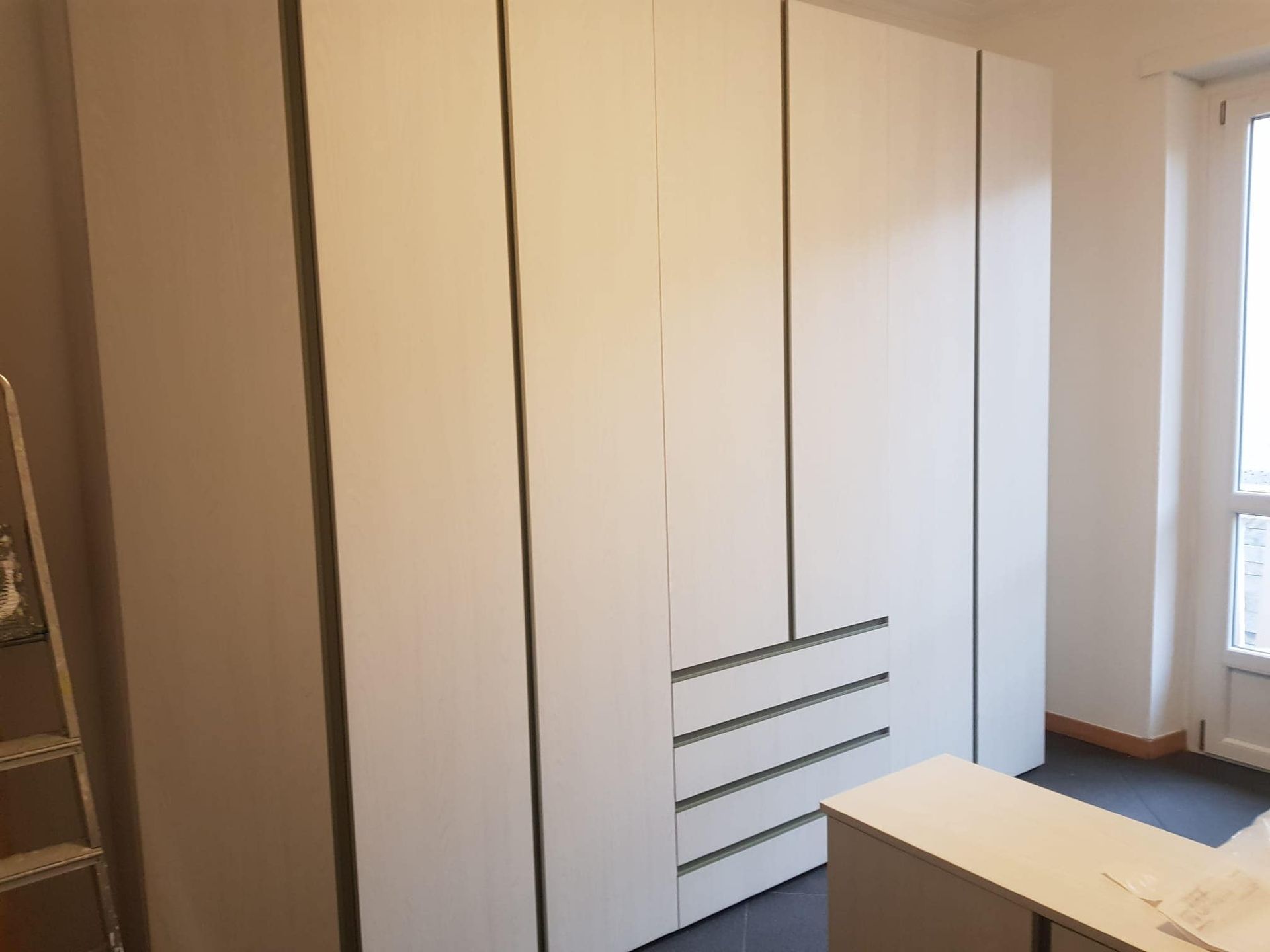 Armadio bianco a muro con tre cassetti, accanto a una scrivania e una finestra in una stanza.