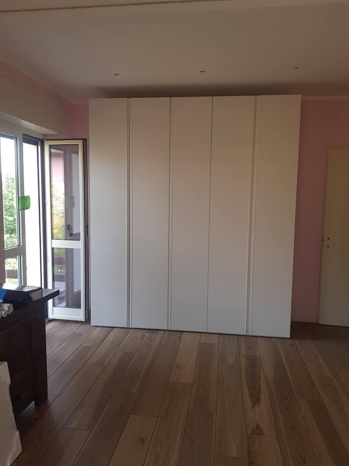 Grande armadio bianco contro una parete rosa in una stanza con pavimento in legno e porta-finestra.