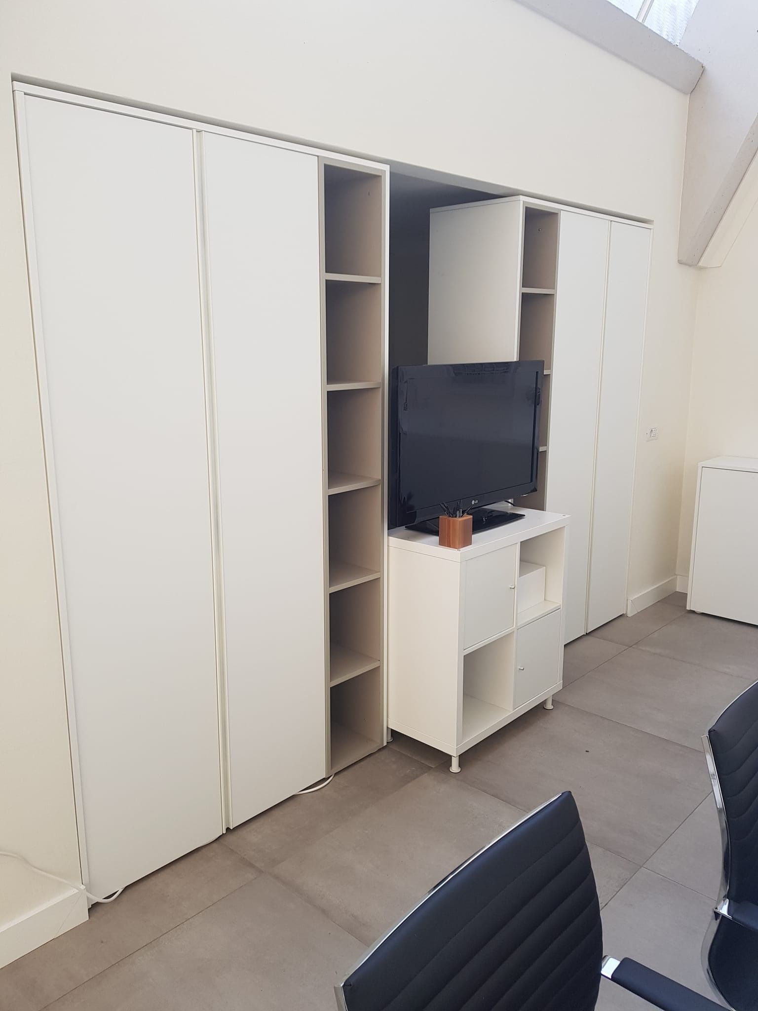 Mobile a parete color crema con ripiani, TV e ante scorrevoli; due sedie nere di fronte.