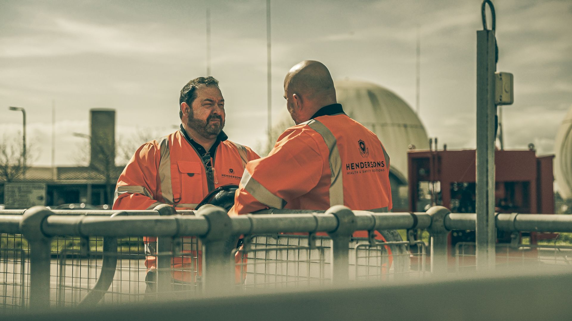 Two men in orange safety vests are standing next to a fence talking to each other.