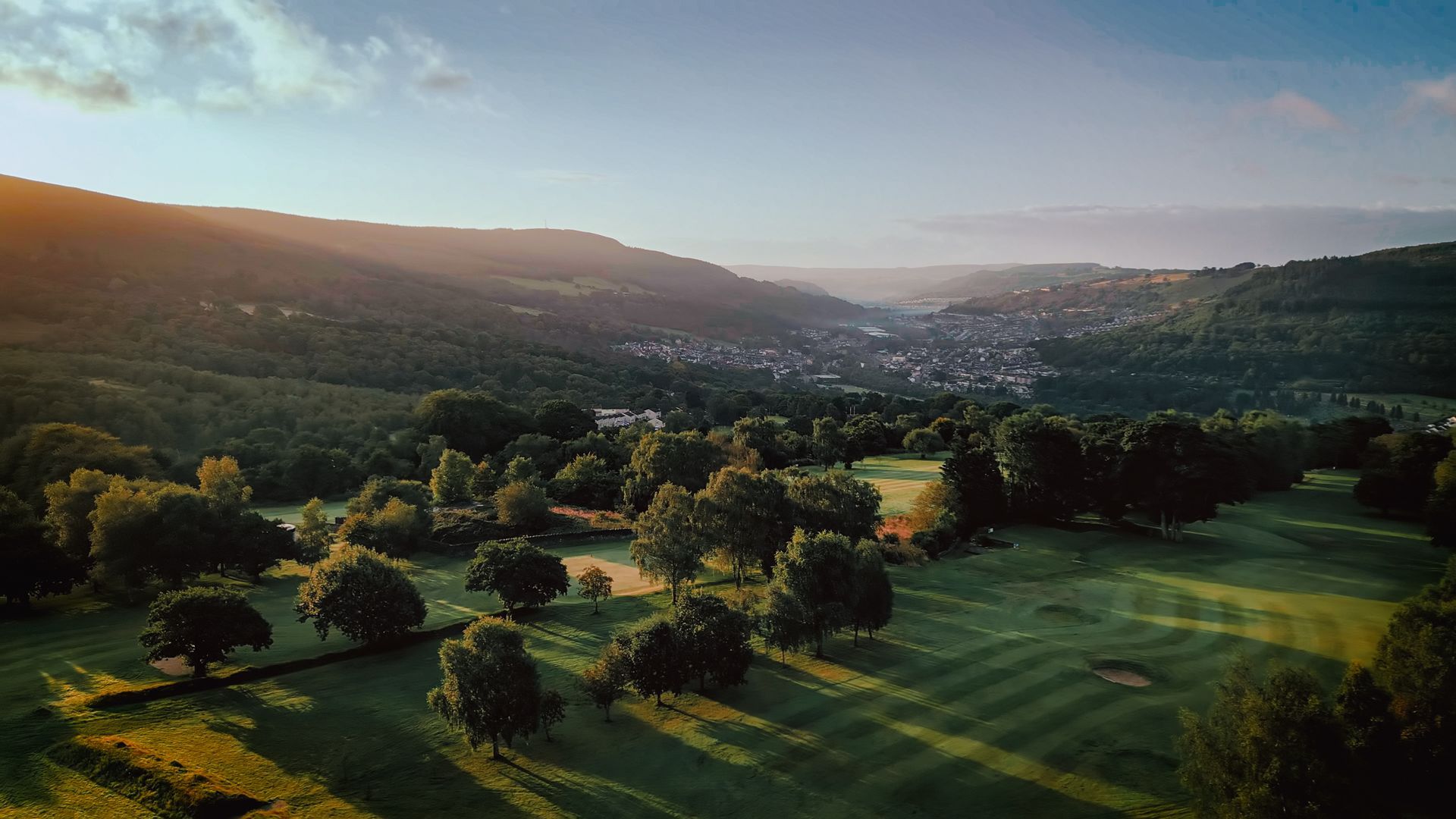 Green golf course nestled in a valley with trees, hills, and a town in the background during a golden sunset.