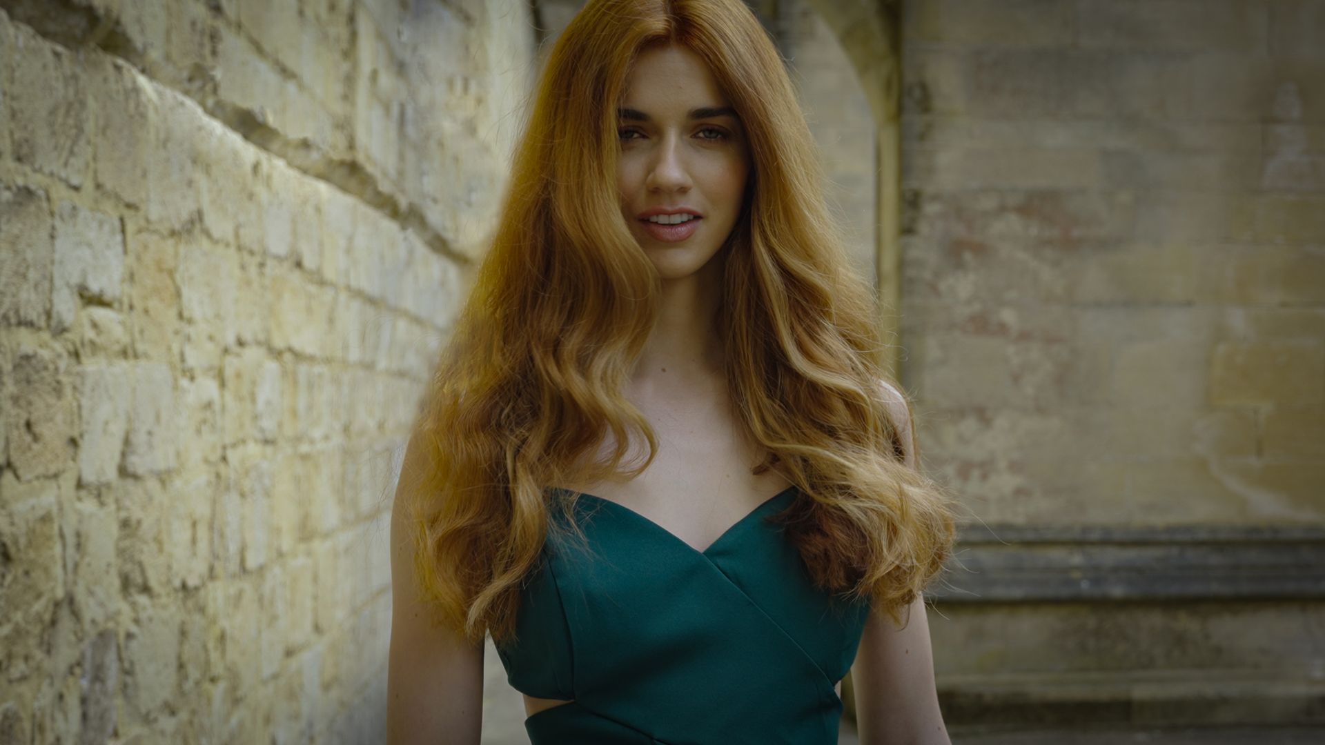 Woman with red hair in green dress near a stone wall, looking towards the camera.