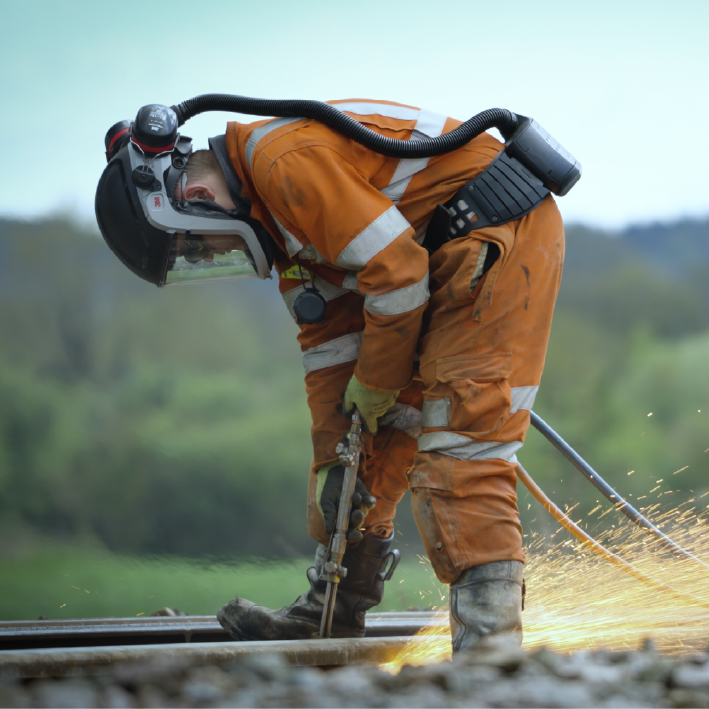 Person in orange protective suit welding on train tracks, sparks flying.