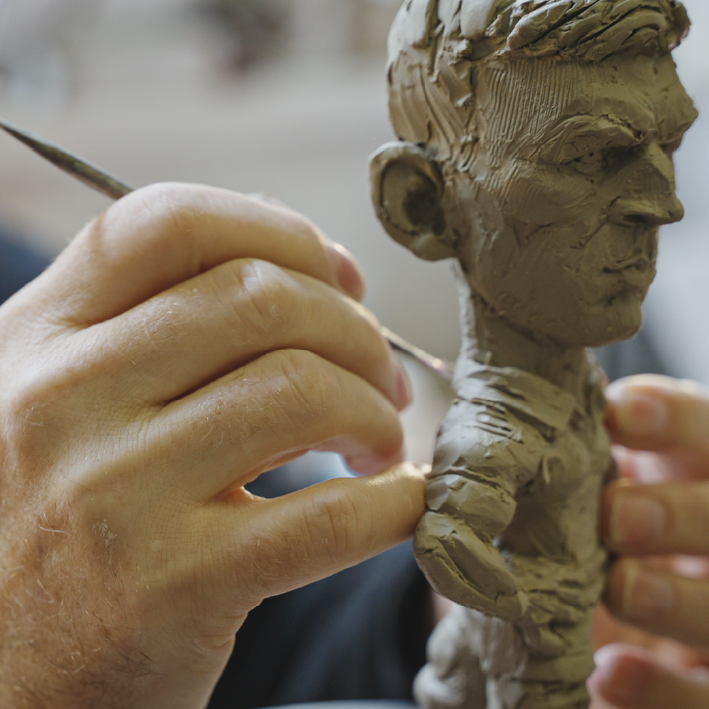 Hands sculpting clay figure of a man with a tool.