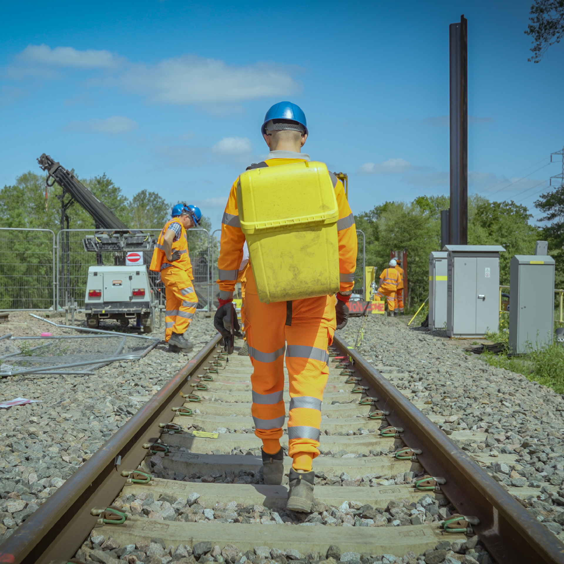 Railway workers in orange jumpsuits walk on tracks with a bright yellow pack on their back.