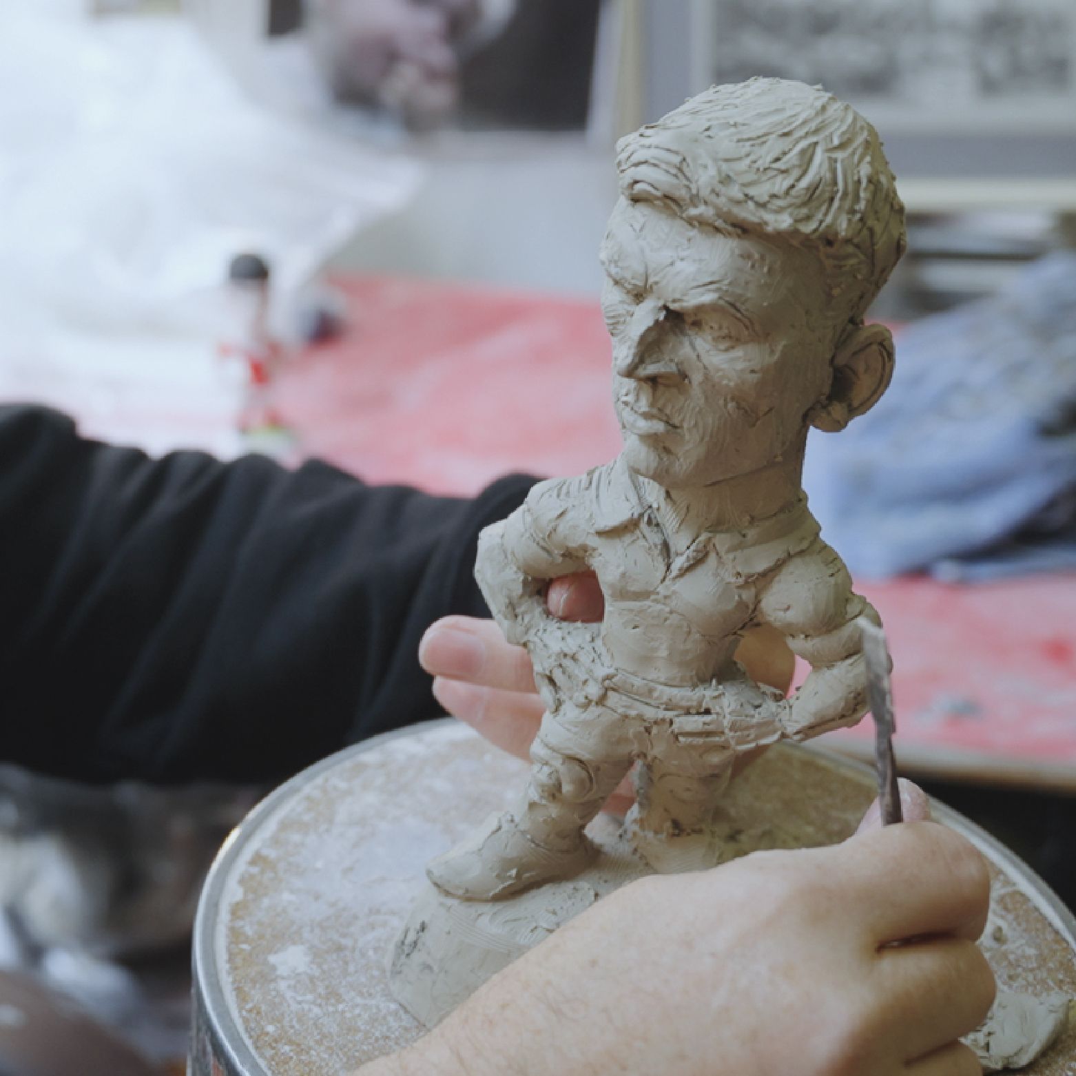 Sculptor working on a clay figurine of a man; indoors with tools, hands, and a workbench.
