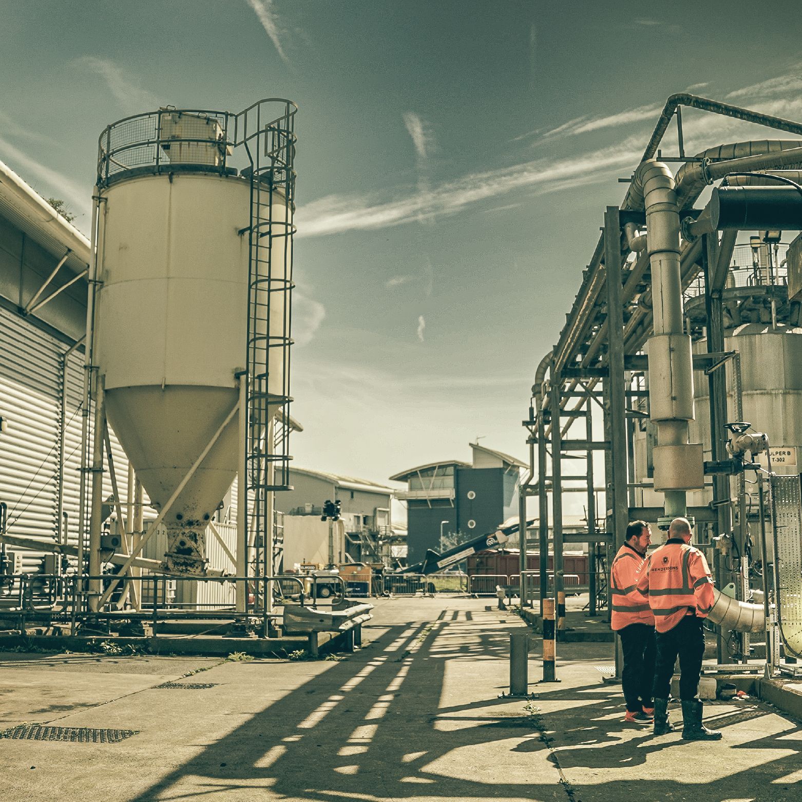 Two workers in safety vests at an industrial site near silos and pipes on a sunny day.
