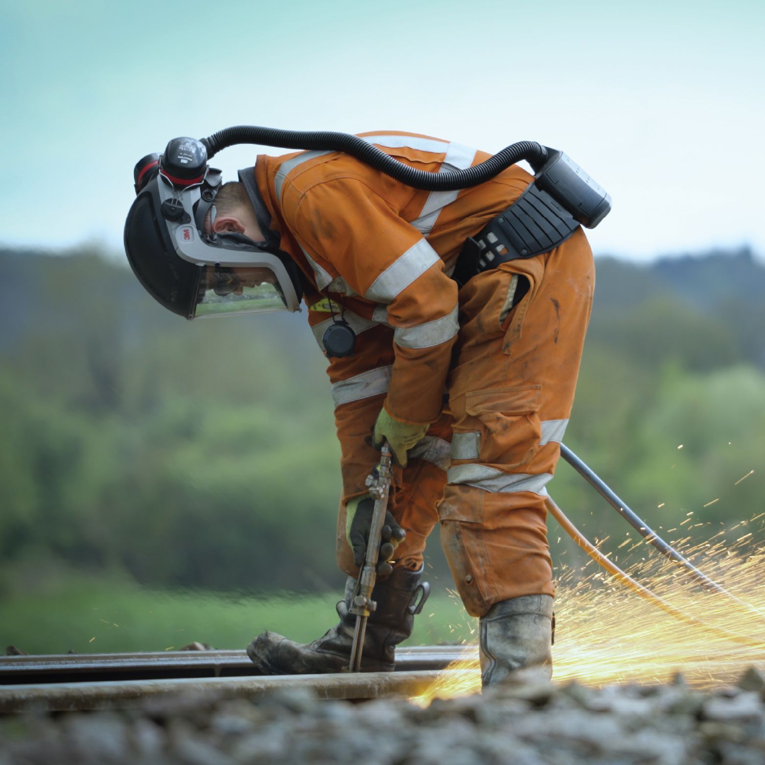 Rail worker in orange suit, helmet, and respirator, cutting track with sparks flying.