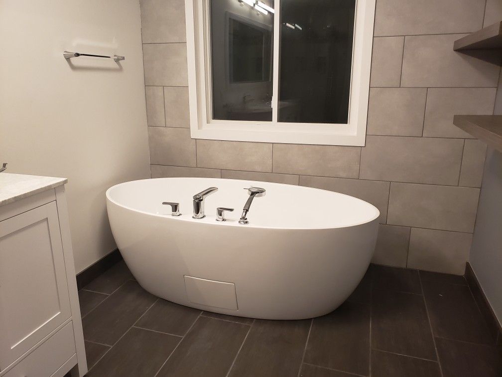 Modern white oval bathtub in a gray-tiled bathroom, near a window and vanity.