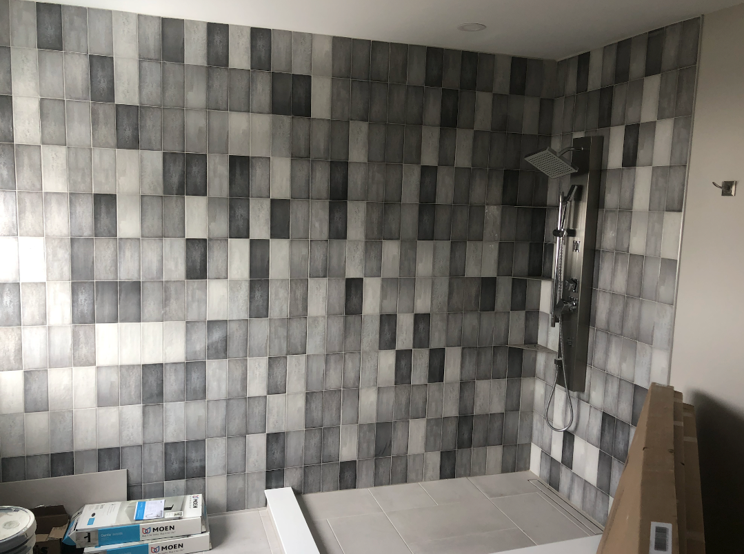 Gray and white tiled shower wall, shower fixtures, and boxes in a room under construction.