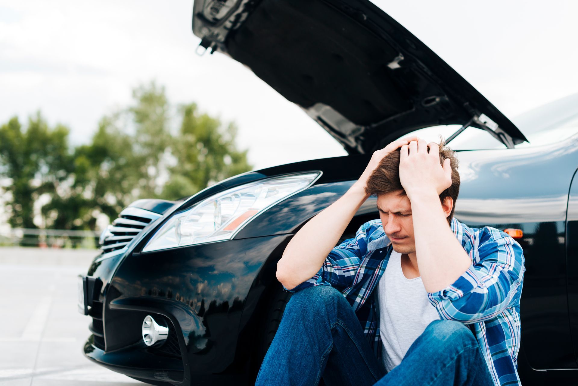 A man is sitting on the ground next to a broken down car with the hood up.