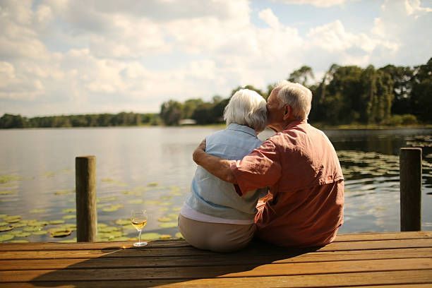 Elderly couple embracing on a dock, overlooking a lake; one holds a glass of wine.